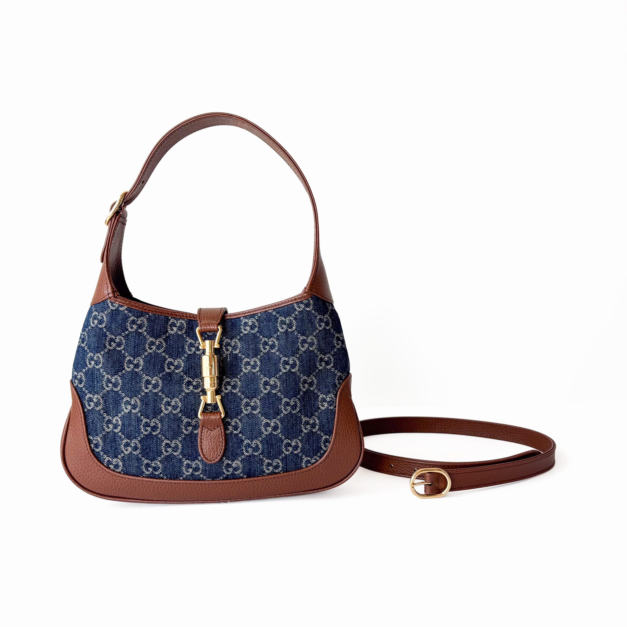 Front view of a small Gucci Jackie 1961 shoulder bag in blue GG monogram denim with brown leather trim, gold-tone piston hardware, and an unattached brown leather extension strap resting on Gucci Jackie 1961 Small Shoulder Bag – GG Monogram Denim Canvas & Brown Grained Leather - Image 1